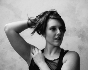  Black and white headshot of Heather (female, early 30s) with one arm across her body resting on shoulder, the other holding her long brown hair behind her head. Her gaze looks off to the side of the camera. Heather wears a black mesh tank top.